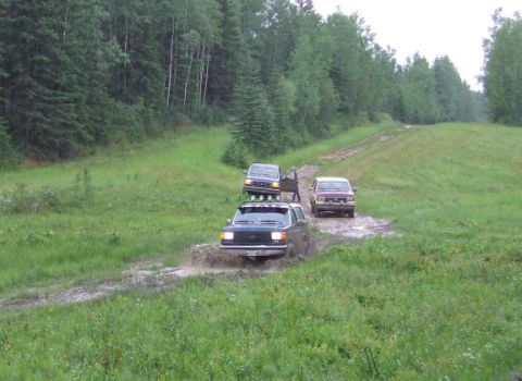 1988 Ford Bronco - Several SUVs navigate a muddy off-road path surrounded by dense forest.