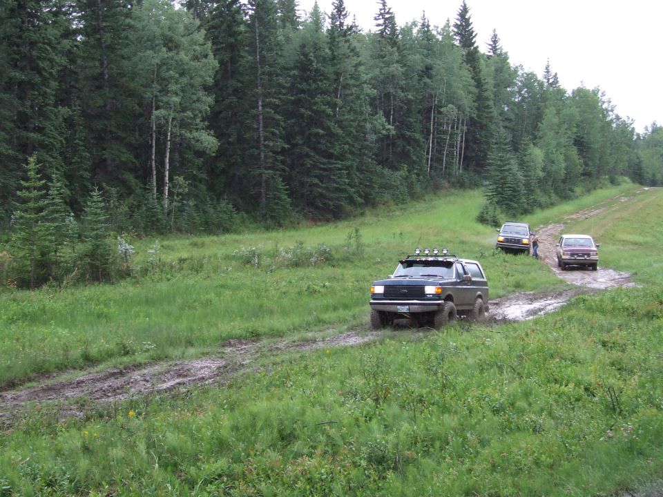 Dark blue Ford Bronco driving through mud on a dirt road with trees in the background