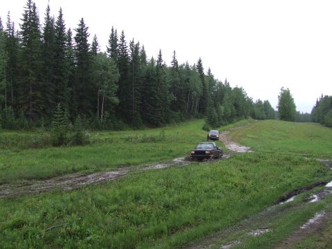 1988 Ford Bronco - Two trucks navigate a wet and muddy path lined with trees under an overcast sky.