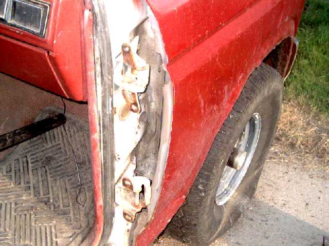 Close-up of a red SUV's open door frame and rusty hinges, showing the interior floor mat and part of the tire