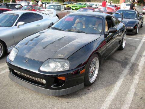 1998 Ford Crown Victoria - black Toyota Supra coupe with a ribbed hood and mesh wheels sits among sports cars at an outdoor meet.