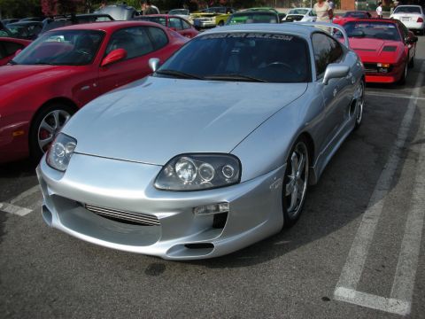 1998 Ford Crown Victoria - silver Toyota Supra fitted with a custom body kit and projector headlights sits among red coupes at a meet.