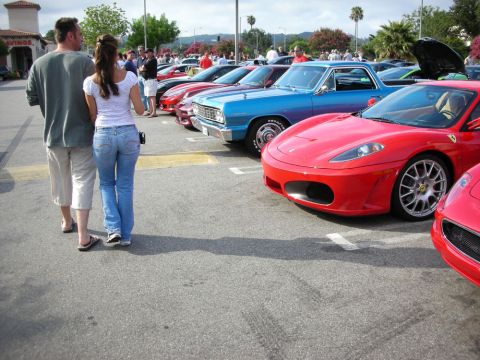 1998 Ford Crown Victoria - couple strolls past a long row of show cars, including a red Ferrari F430 and a blue classic, under a clear sky.
