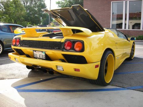 1998 Ford Crown Victoria - yellow Lamborghini Diablo roadster sits with rear engine cover up and a vanity plate in a handicap-marked spot.