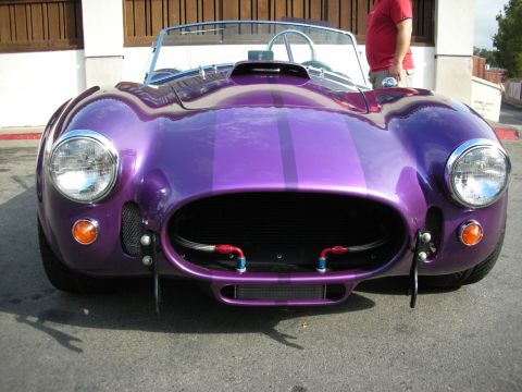 1998 Ford Crown Victoria - purple Shelby Cobra-style roadster with twin racing stripes and chrome headlights sits in a sunlit lot, a man nearby.