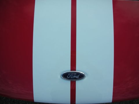 1998 Ford Crown Victoria - Close-up of a red hood with a wide white racing stripe and a chrome Ford oval badge at center.
