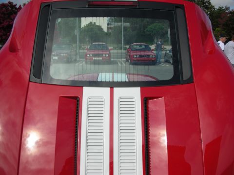 1998 Ford Crown Victoria - Rear deck of a red car with twin white stripes and a louvered rear window reflecting other cars in a lot.