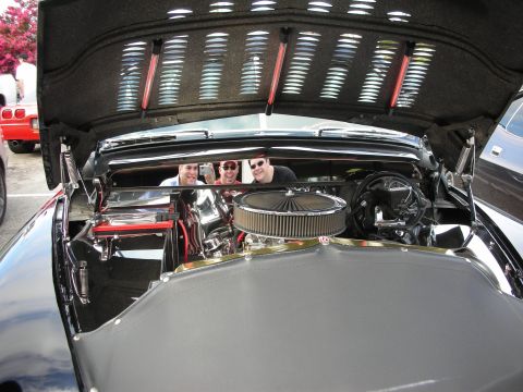 1998 Ford Crown Victoria - Engine bay of a black classic roadster showing a chrome air cleaner and polished components, with three onlookers reflected in the chrome a…