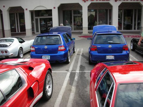 1998 Ford Crown Victoria - Two blue Volkswagen Golf hatchbacks parked rear-to-front flanked by red sports cars in front of a storefront at a car meet.