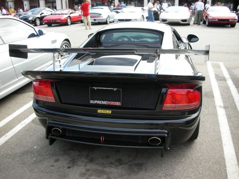 1998 Ford Crown Victoria - Rear of a black mid-engine sports car with a tall rear wing, dual exhaust and a SUPREME POWER license plate among cars at a show.