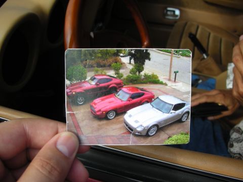 1998 Ford Crown Victoria - hand holds a small printed photo showing three Datsun Z-style sports cars, red and silver, parked in a driveway, taken inside a car.