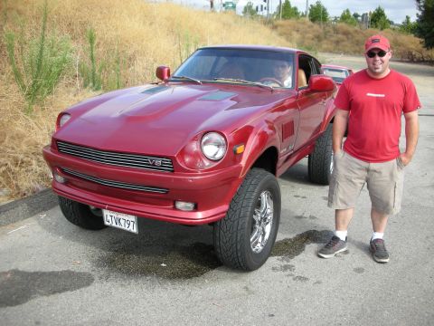 1998 Ford Crown Victoria - man in a red shirt stands beside a lifted red Datsun Z-style coupe with a V8 grille badge and chrome wheels on a roadside.