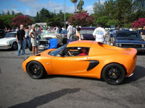 1998 Ford Crown Victoria - orange Lotus Elise sits in profile on black wheels as a crowd browses classic cars in the background.