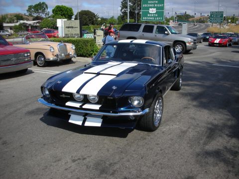 1998 Ford Crown Victoria - dark blue Shelby-style Mustang fastback with white racing stripes parked near a freeway sign at a car show.