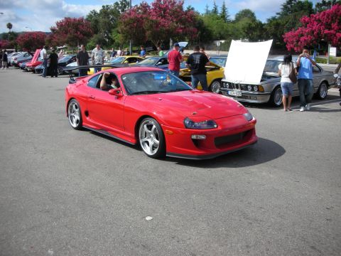 1998 Ford Crown Victoria - red Toyota Supra in front three-quarter view on polished multi-spoke wheels, rolling through a car-show lot.