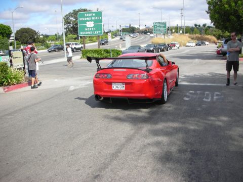 1998 Ford Crown Victoria - red Toyota Supra with a tall rear wing sits at a stop-marked intersection, plate reading KING-ERM.