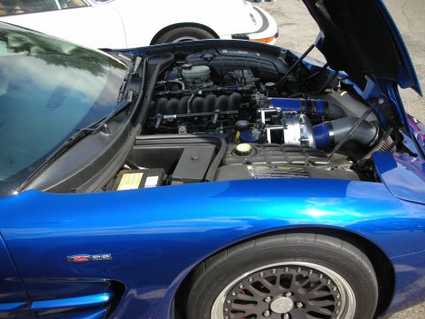 1998 Ford Crown Victoria - Engine bay of a blue Corvette Z06 with blue intake piping, seen over the front fender and a black multi-spoke wheel.