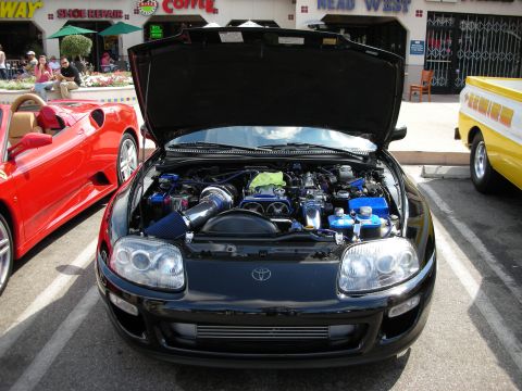 1998 Ford Crown Victoria - black Toyota Supra with its hood raised, displaying the inline-six engine and blue accents, beside a red Ferrari at a show.