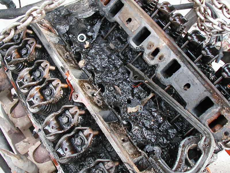 V8 engine with valve covers off, rocker arms and lifter valley caked in thick black oil sludge, hanging from chains