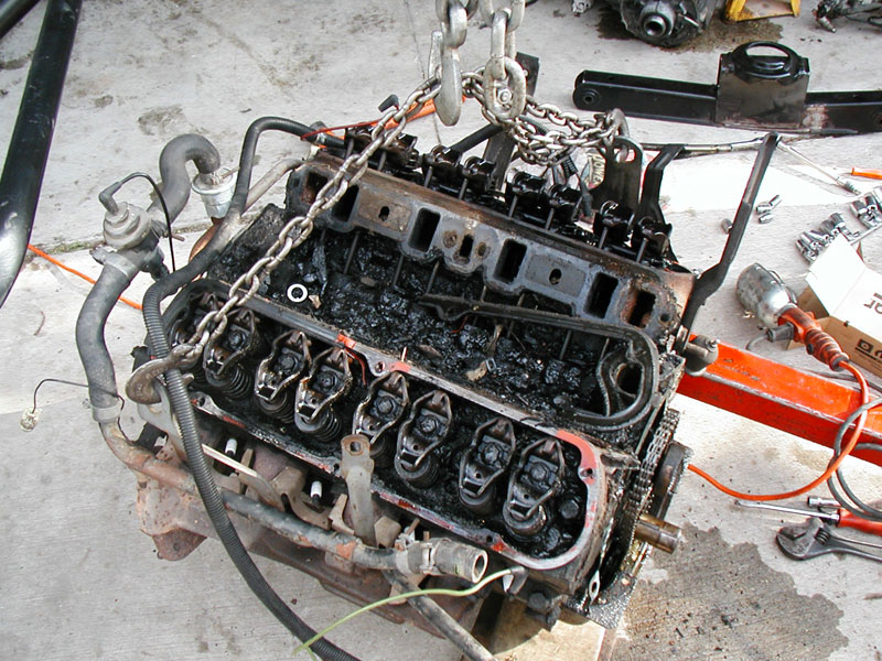 Removed V8 engine on a lifting chain with valve covers off, showing heavy black sludge buildup on rockers and valley