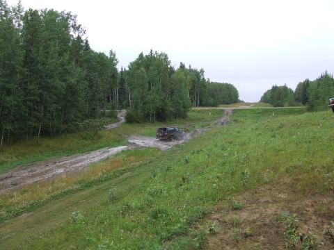 1988 Ford Bronco - A dark SUV navigates a muddy track through a wooded area, with another vehicle visible in the distance.