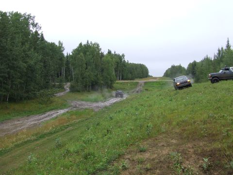 1988 Ford Bronco - Multiple SUVs navigate a wet and muddy off-road course surrounded by trees under an overcast sky.