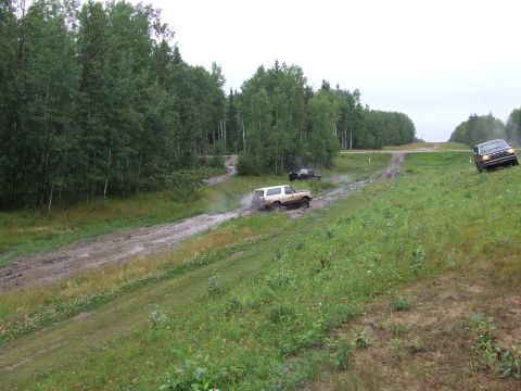 1988 Ford Bronco - Three SUVs navigate a challenging muddy off-road course with dense trees lining the path.
