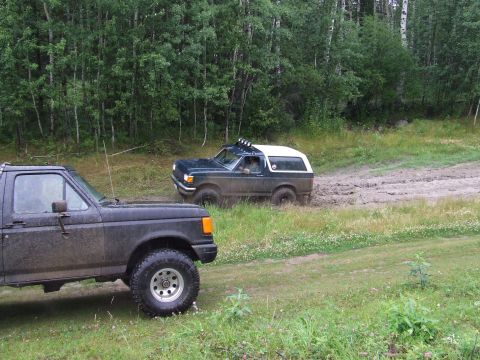 1988 Ford Bronco - A dark green 1988 Ford Bronco with a white top navigates a muddy track, while another dark SUV is parked in the foreground.