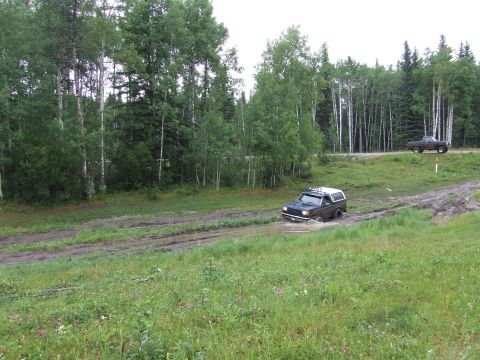 1988 Ford Bronco - A dark SUV with a white top navigates a muddy track, with another vehicle visible on a higher road in the background.