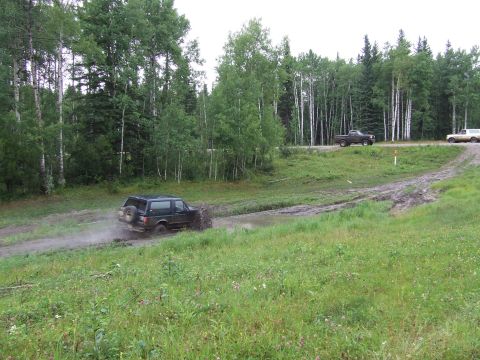 1988 Ford Bronco - A dark 1988 Ford Bronco navigates a muddy track with trees in the background and two other vehicles on a dirt road.