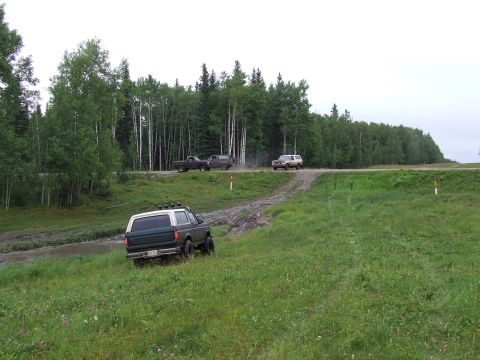 1988 Ford Bronco - A dark green and white Ford Bronco navigates a muddy track off-road, with other vehicles visible on a distant road.