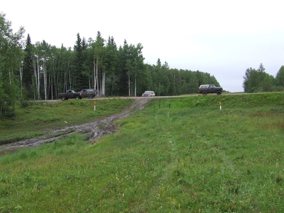 three SUVs driving on a dirt road next to a forest, one vehicle is on a muddy track