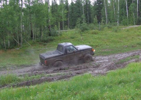 1988 Ford Bronco - A dark-colored Ford Bronco II is driving through a muddy, off-road trail surrounded by trees.