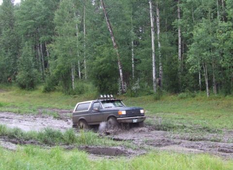 1988 Ford Bronco - A Ford Bronco navigates a challenging muddy path surrounded by trees.