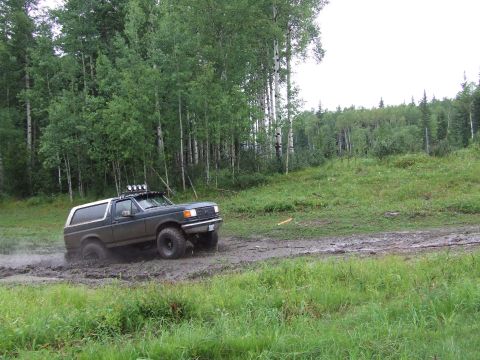 1988 Ford Bronco - A dark green Ford Bronco navigates a muddy trail surrounded by trees.