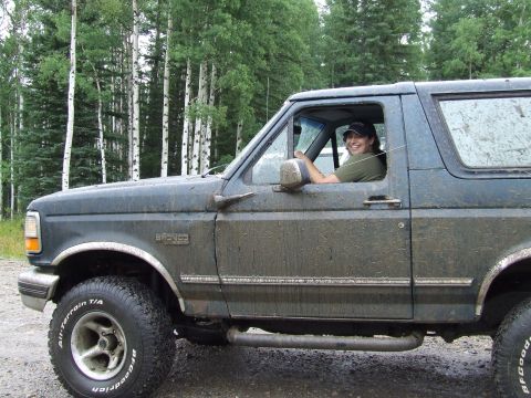 1988 Ford Bronco - Driver in a muddy 1988 Ford Bronco parked on a dirt road with trees in the background.