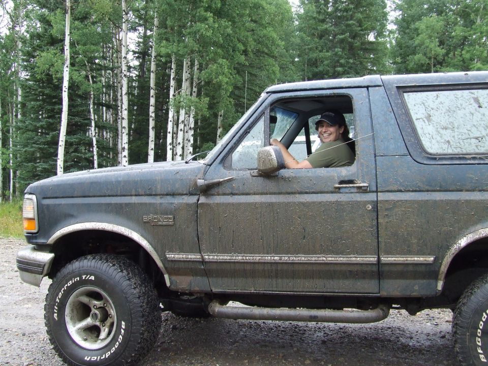1988 Ford Bronco side view, driver smiling, muddy exterior, forest background