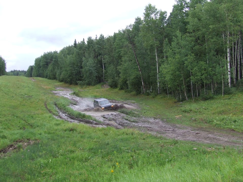 Dark SUV driving through mud on a dirt track, surrounded by trees