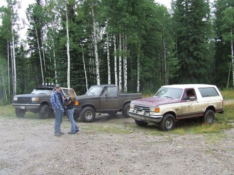 1988 Ford Bronco - Three Ford trucks are parked on a gravel lot with a wooded area in the background.