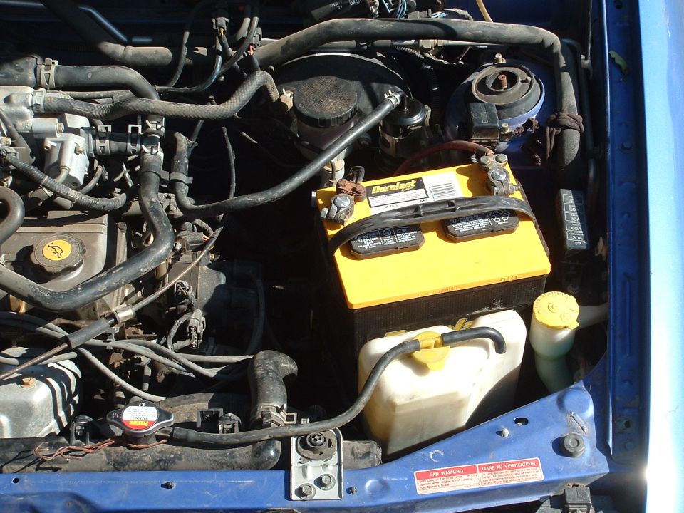 Engine bay with yellow Duralast battery, white fluid reservoir, hoses, and fan warning sticker in a blue car
