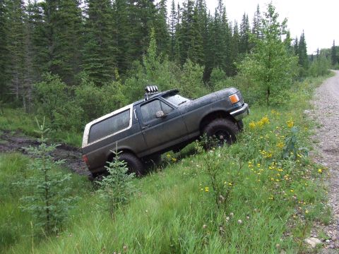 1988 Ford Bronco - A dark green SUV with a white top navigates a muddy, grassy incline next to a dirt road.