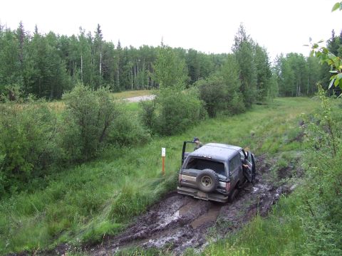 1988 Ford Bronco - A dark SUV is stuck in a muddy trail surrounded by trees, with its doors open.