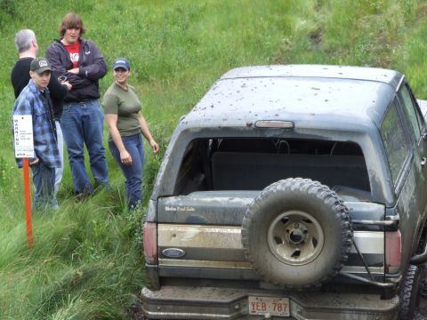 1988 Ford Bronco - A Ford Bronco is partially submerged in mud, with several people standing nearby on grassy terrain.