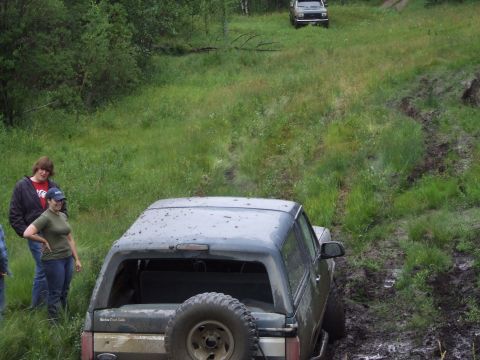 1988 Ford Bronco - A dark 1988 Ford Bronco navigates a muddy off-road trail, with two onlookers observing its progress.