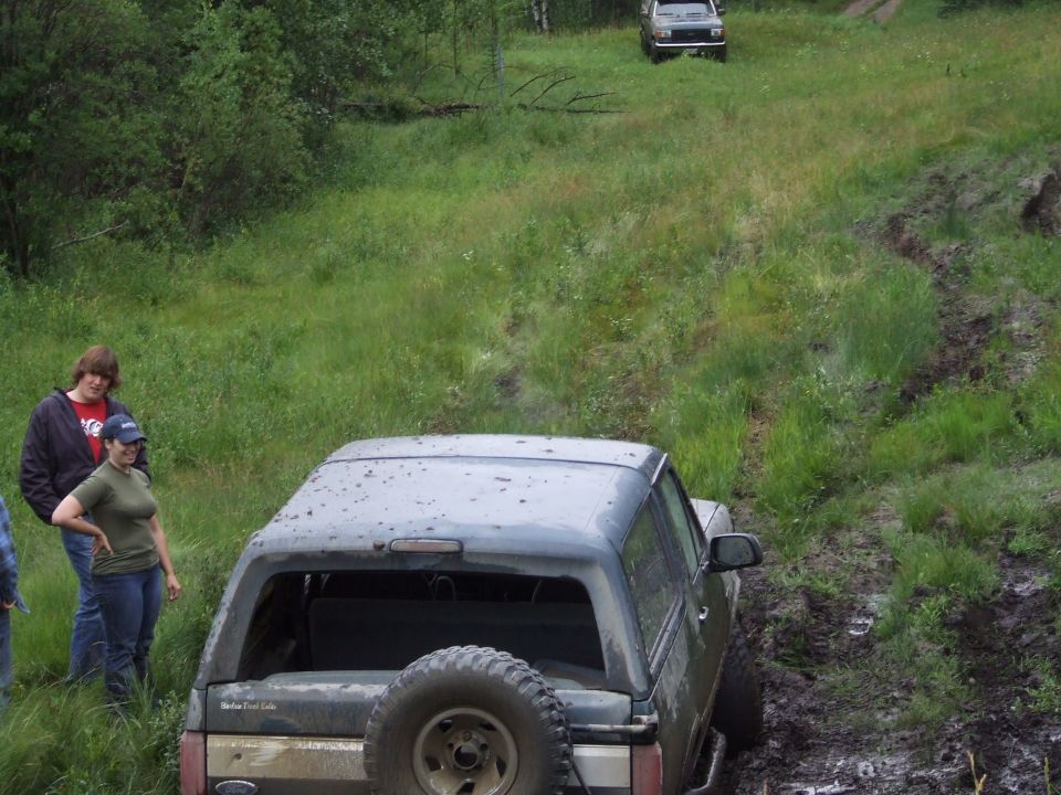 Dark 1988 Ford Bronco stuck in mud, rear view. Two people stand nearby