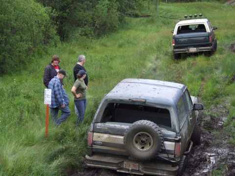 1988 Ford Bronco - Two vehicles navigate a muddy off-road course with onlookers.