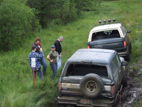 1988 Ford Bronco - Two mud-splattered full-size Ford Broncos sit in a rutted mud track on a grassy trail while four people stand in the tall grass beside an o…