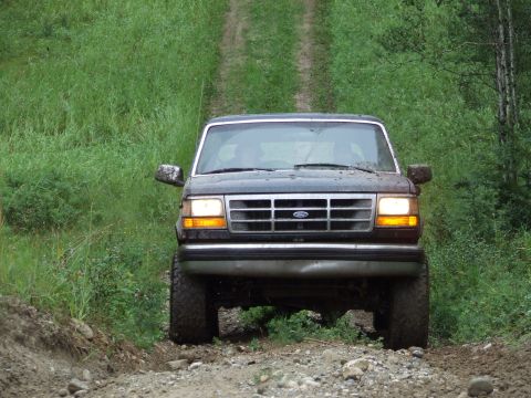 1988 Ford Bronco - This dark-colored Ford Bronco is navigating a rough, unpaved path surrounded by lush green vegetation.