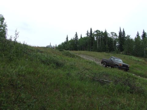 1988 Ford Bronco - Two trucks navigate a dirt road on a grassy incline with trees in the background.