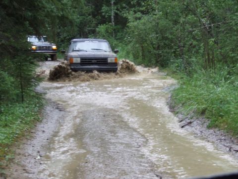 1988 Ford Bronco - Two SUVs navigate a water-filled dirt road, kicking up spray as they drive through the wooded area.